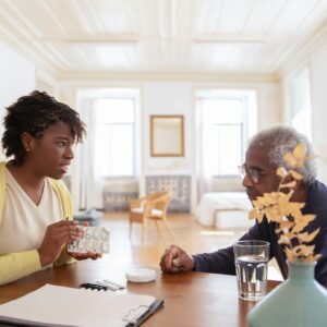 An elderly man sits with a caregiver discussing medication at a table in a bright room.